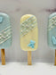 Three decorative popsicle-shaped cakes with blue and green designs on a gray background