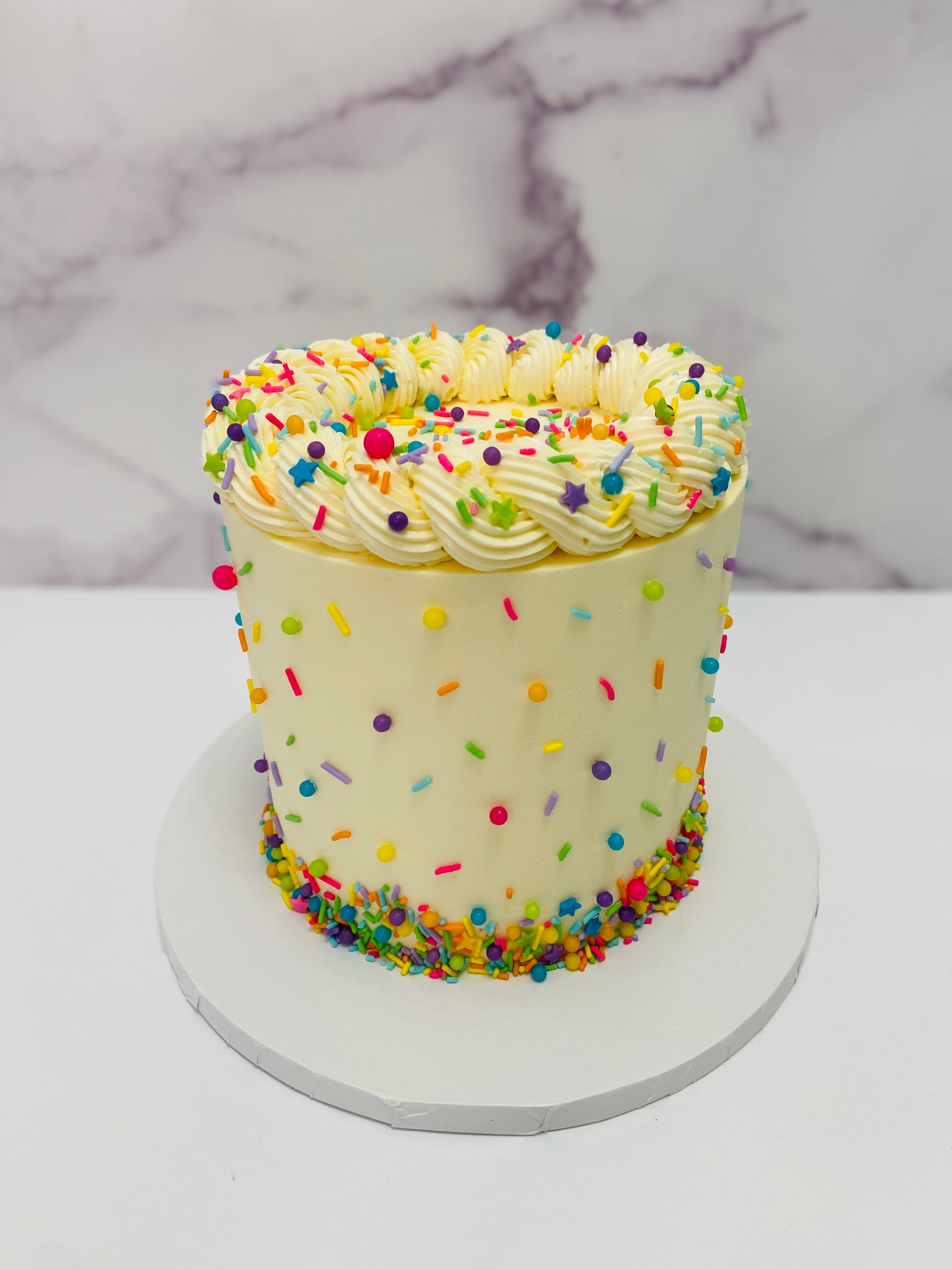 Small round cake with white frosting and colorful sprinkles on a marble background