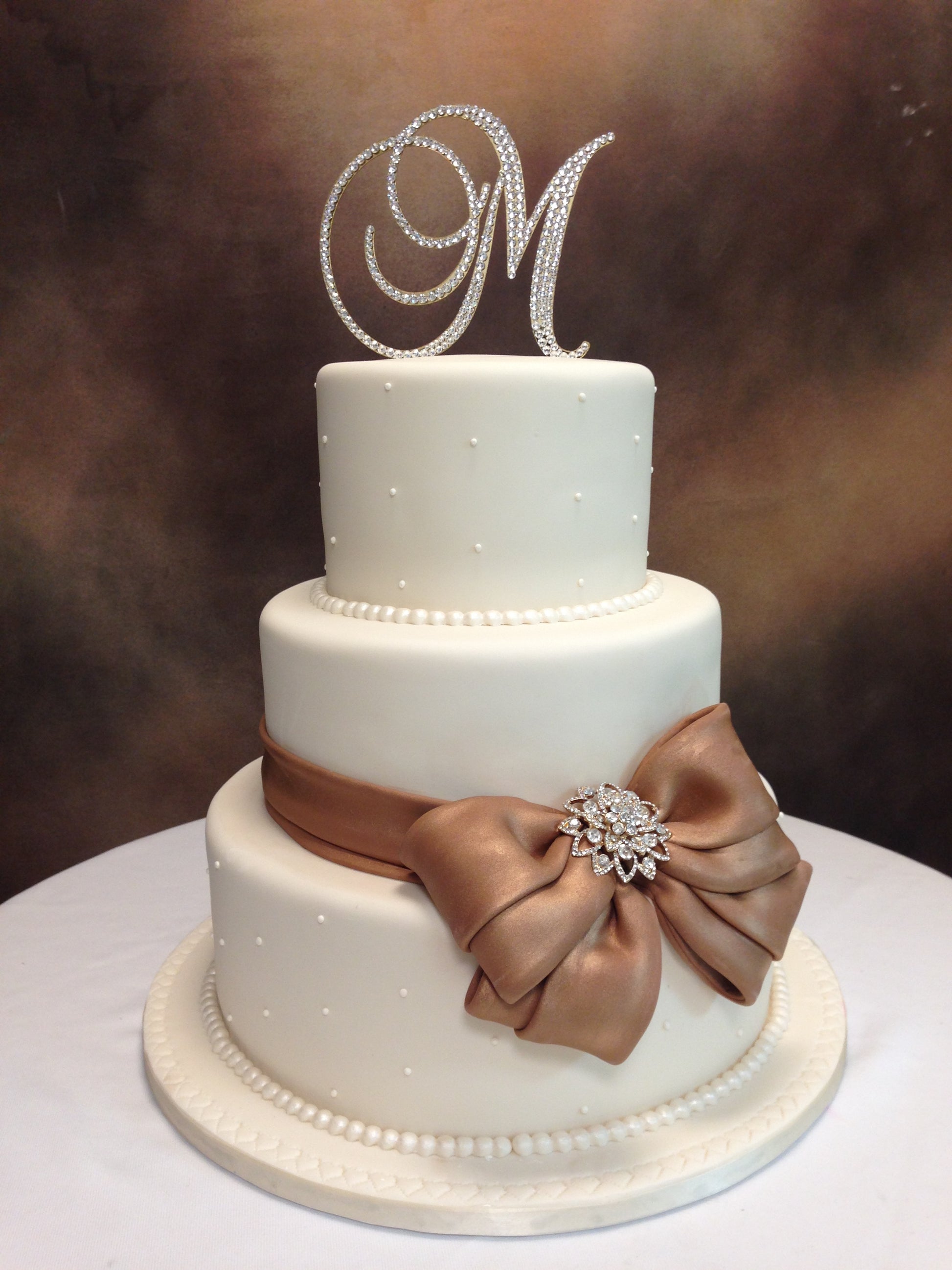 Three-tiered wedding cake with a decorative bow and 'M' monogram on a blurred background