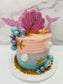 Mermaid-themed birthday cake with pink and blue colors, seashell decorations, and pearls on a white marble background.