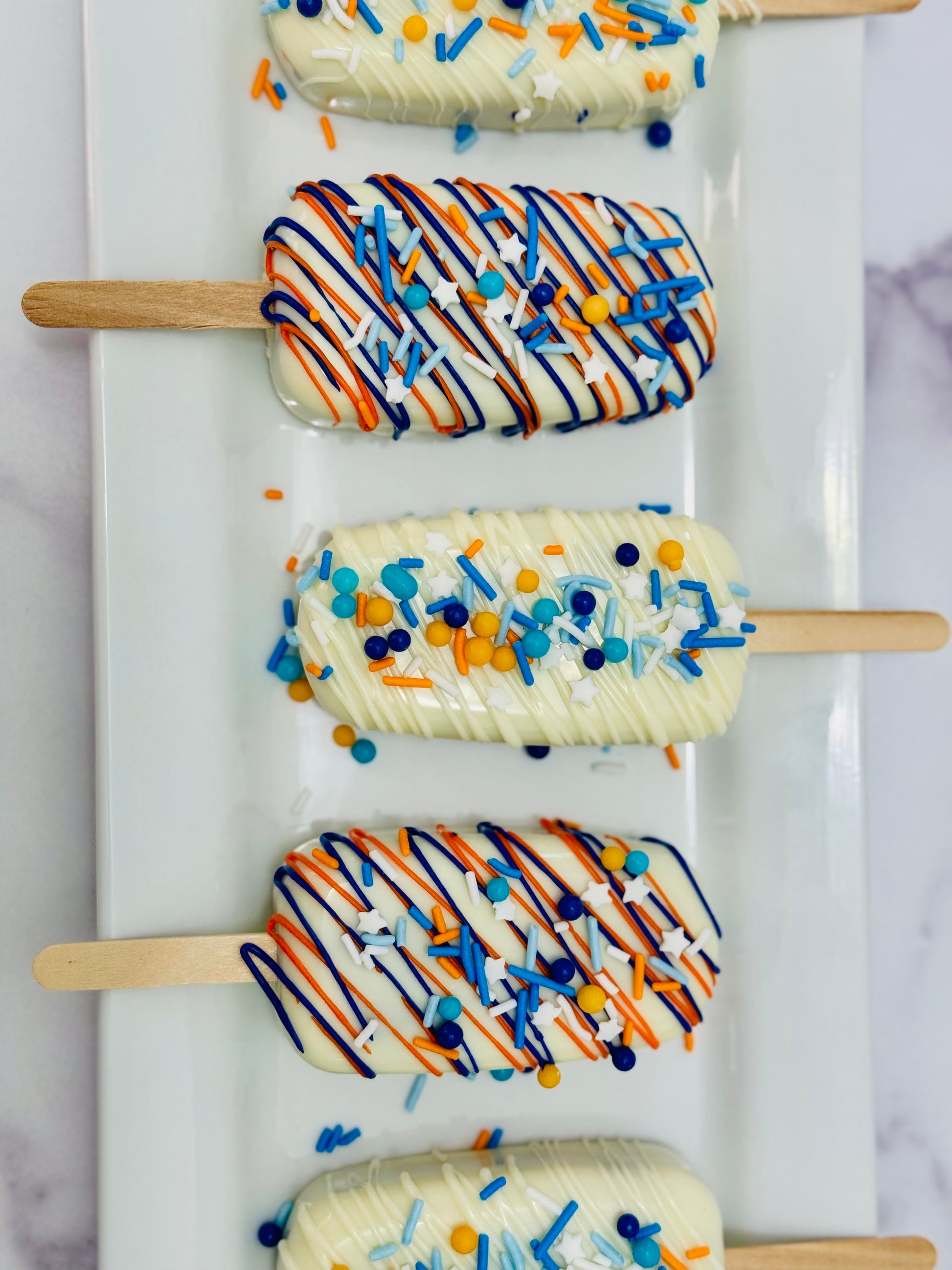 OKC Thunder Basketball Cakesicles with white chocolate drizzle and colorful sprinkles on wooden sticks.