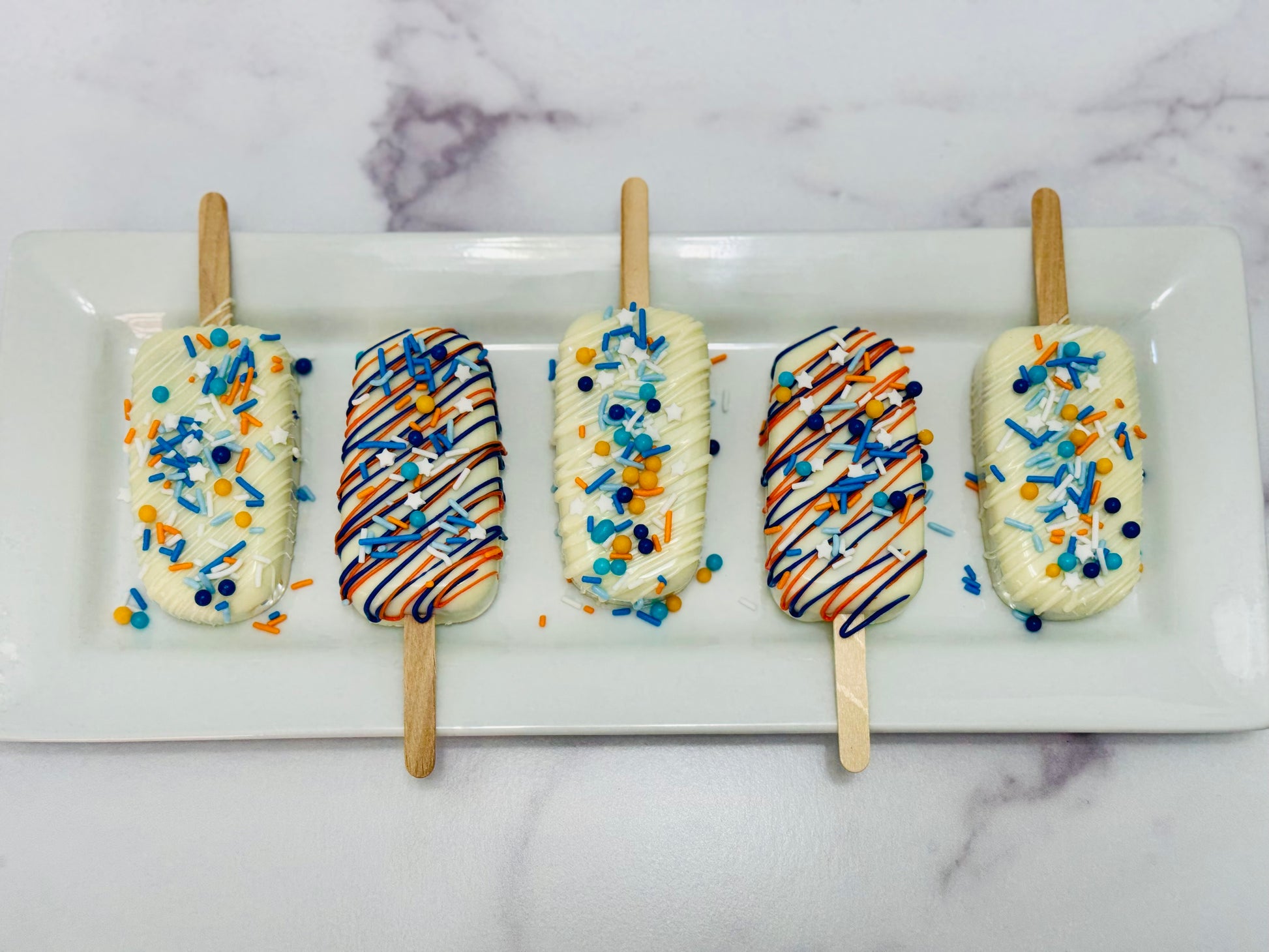 Five decorated OKC Thunder Basketball Cakesicles on sticks with colorful toppings on a white plate.