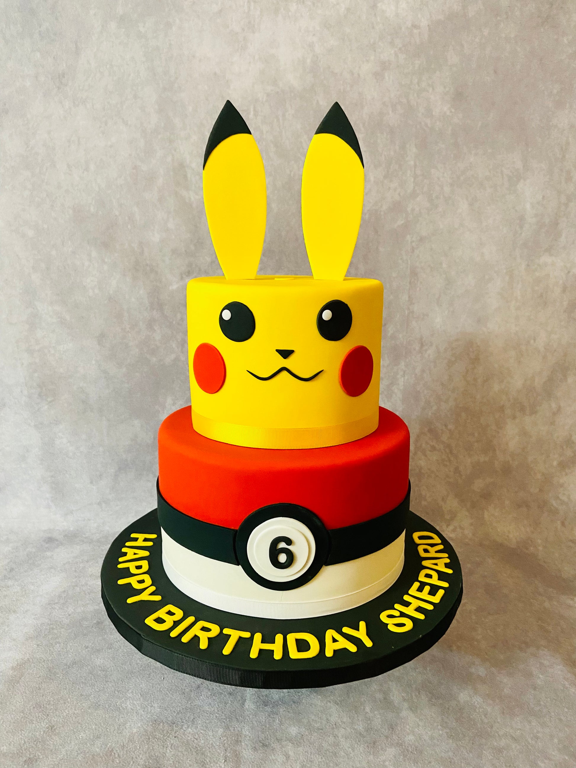 Two-tiered birthday cake designed like Pikachu with a Poké Ball, on a neutral background.