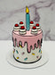 Small birthday cake with pink drip design, colorful sprinkles, and a single candle on a marble background.