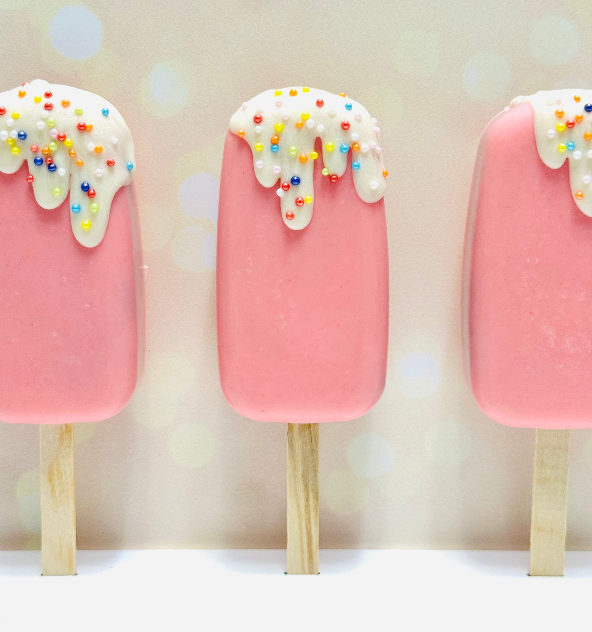 Three pink popsicles with white drizzle and colorful sprinkles on a light background