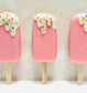 Three pink popsicles with white drizzle and colorful sprinkles on a light background