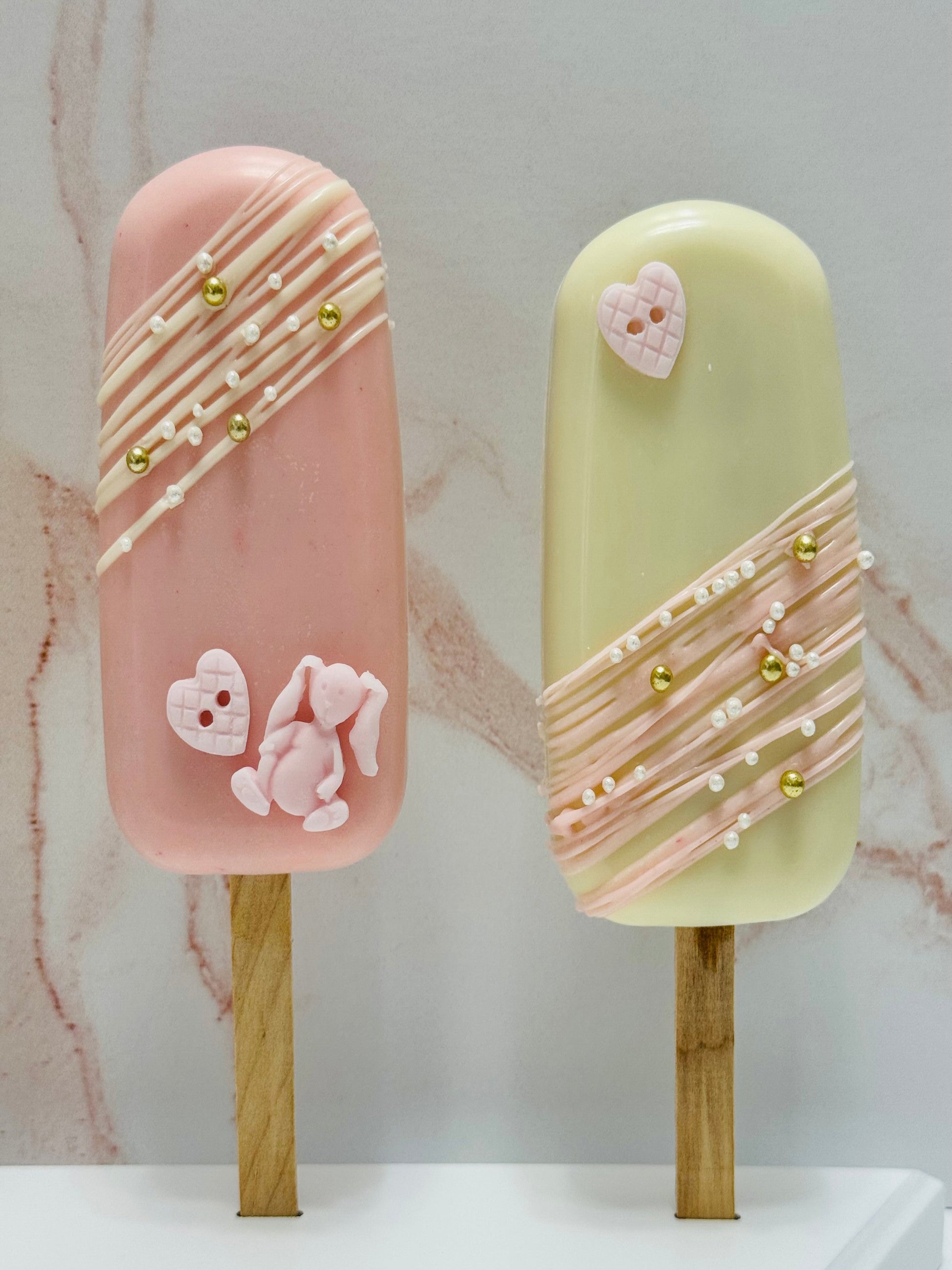 Two decorative popsicle-shaped cakes on sticks with pink and green colors and decorative elements.